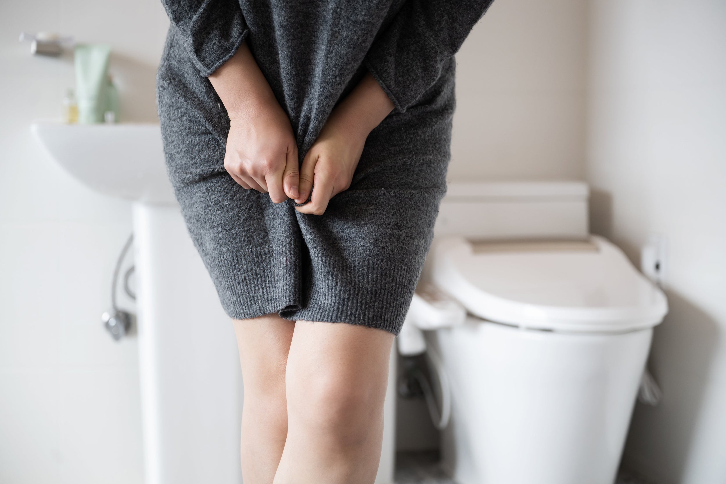 Eine Frau steht im Badezimmer und haelt sich das Kleid zusammen, im Hintergrund Waschbecken und Toilette – Harndrang bei Harninkontinenz.
