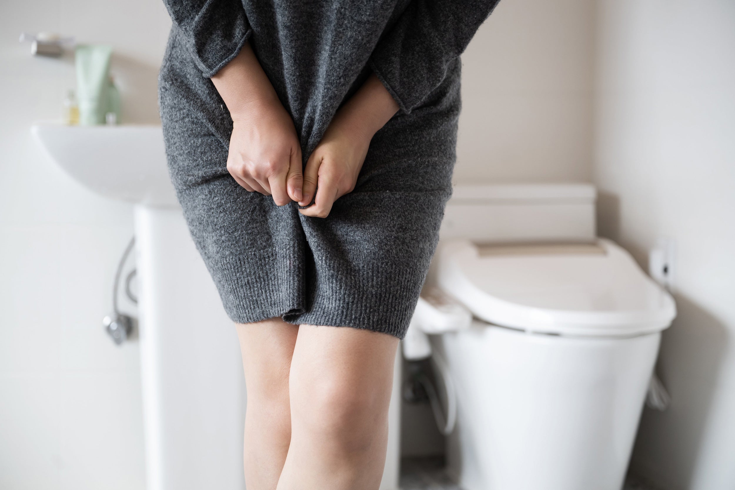 Eine Frau steht im Badezimmer und haelt sich das Kleid zusammen, im Hintergrund Waschbecken und Toilette – Harndrang bei Harninkontinenz.