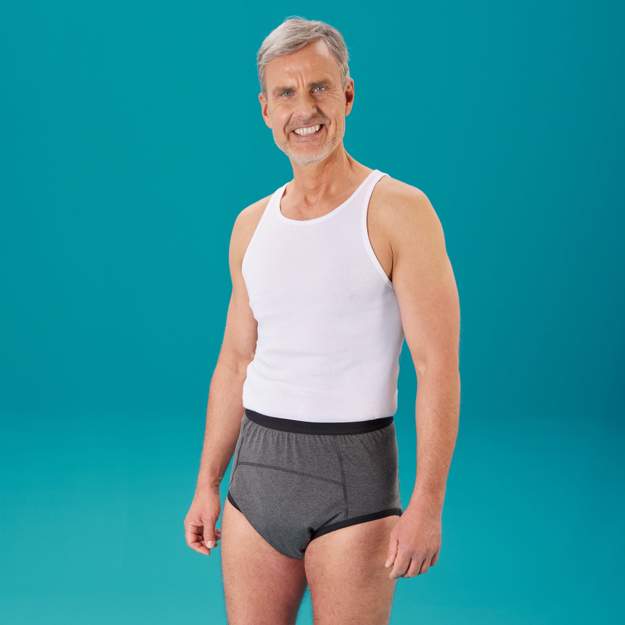 Ein mittelalter Mann mit blauen Augen, grauem Haar und Drei-Tage-Bart trägt den suprima Inkontinenz-Bodyguard-Slip 3 1259 in grau und ein weißes Tanktop. Er steht vor grünem Hintergrund und lächelt in die Kamera.
