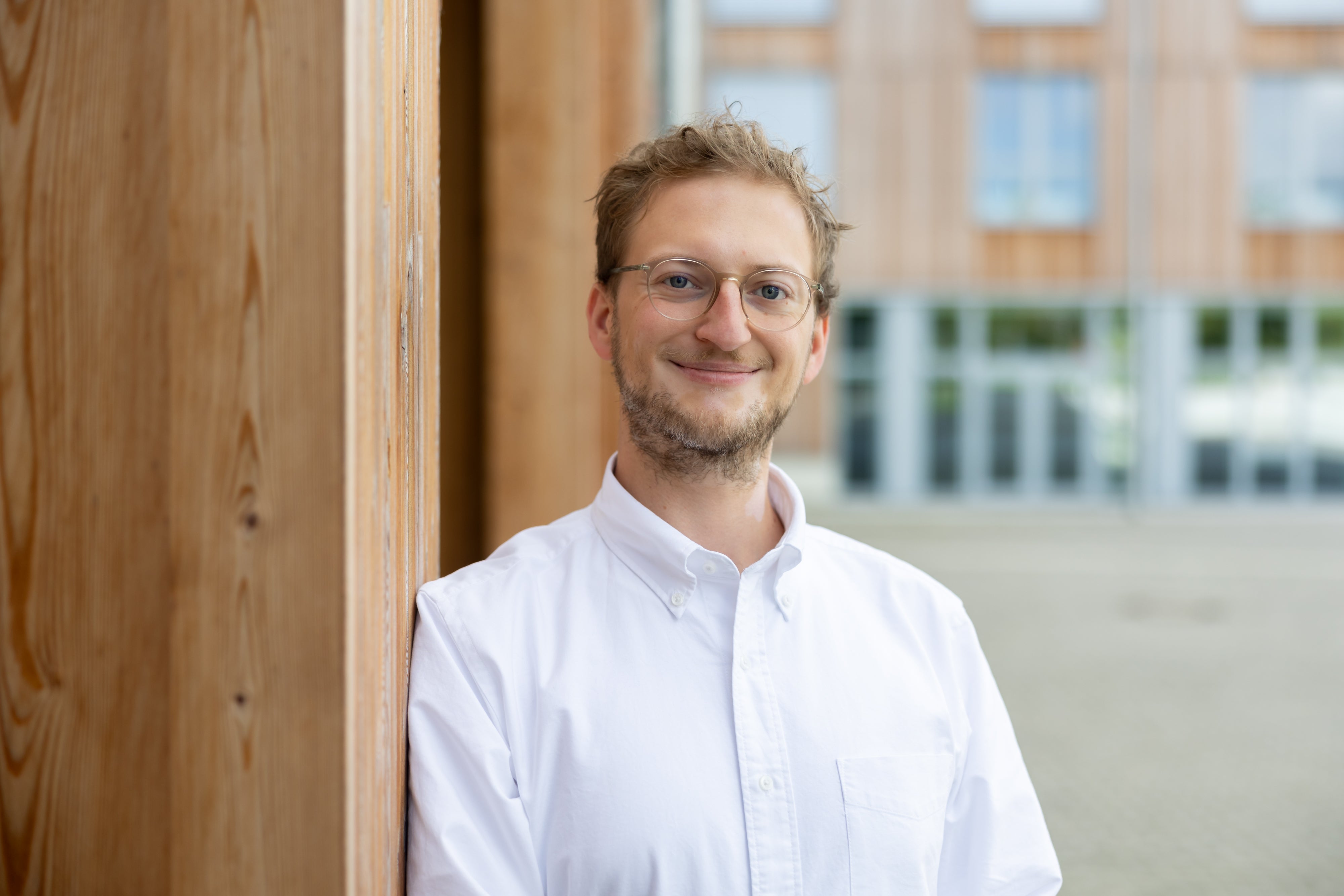 Portrait von David Richter, Geschaeftsfuehrung suprima GmbH, lachend vor einem Holzpfeiler und Gebaeude im Hintergrund