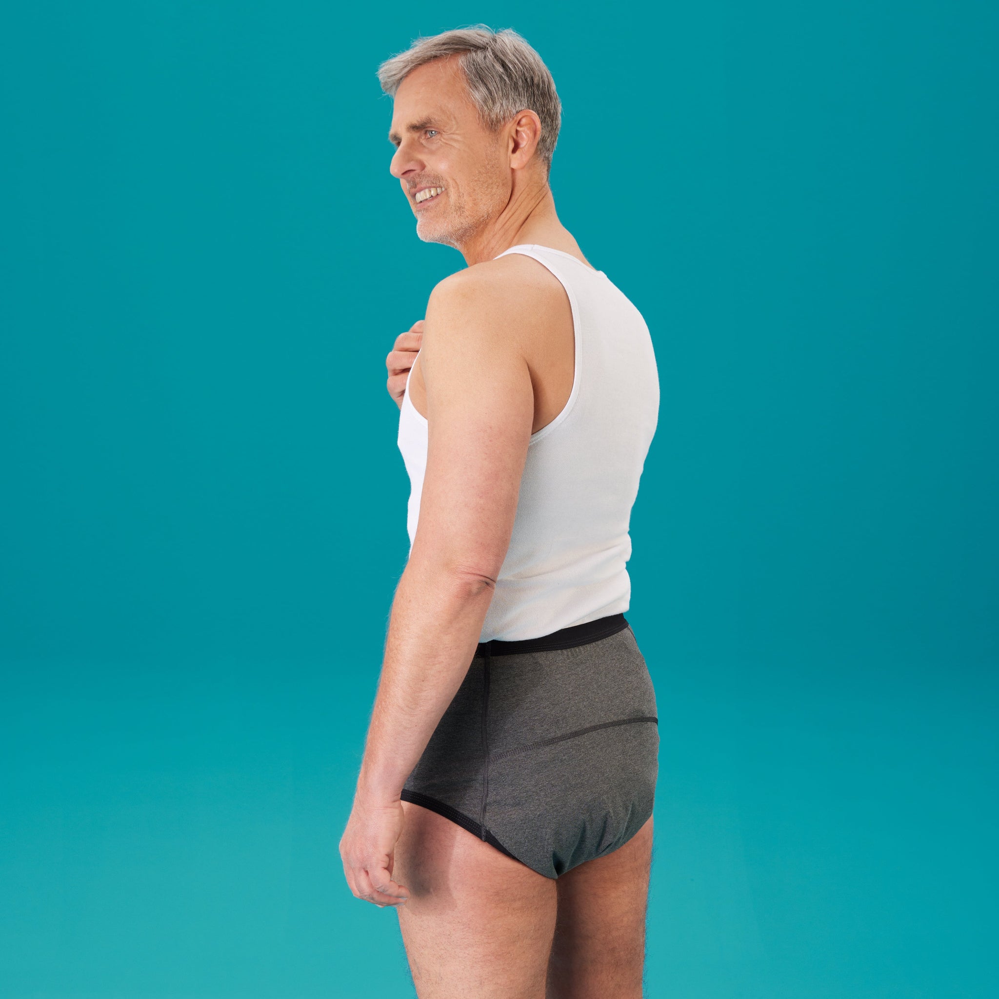 Ein mittelalter Mann mit blauen Augen, grauem Haar und Drei-Tage-Bart trägt den suprima Inkontinenz-Bodyguard-Slip 3 1259 in grau und ein weißes Tanktop. Er steht mit dem Rücken zur Kamera vor grünem Hintergrund. 